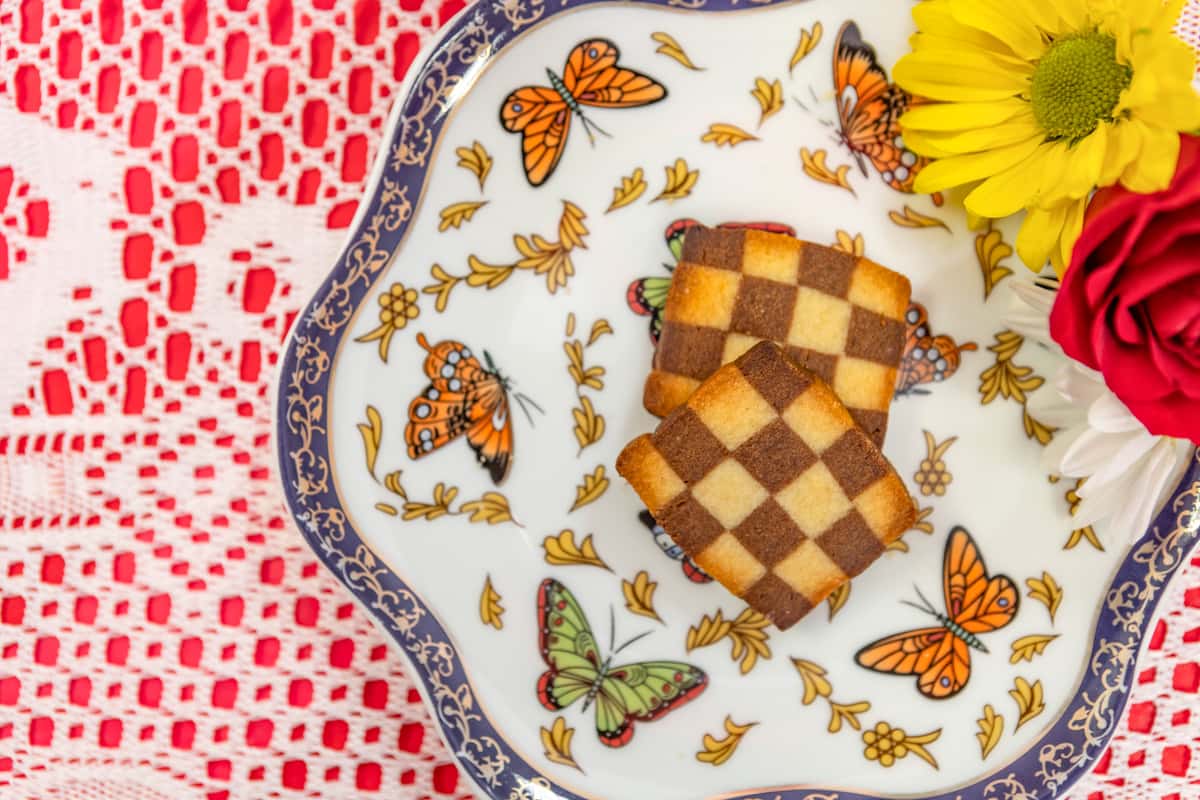 Checkerboard shortbread cookies with vanilla and chocolate squares, served on a butterfly-themed plate with floral garnish.