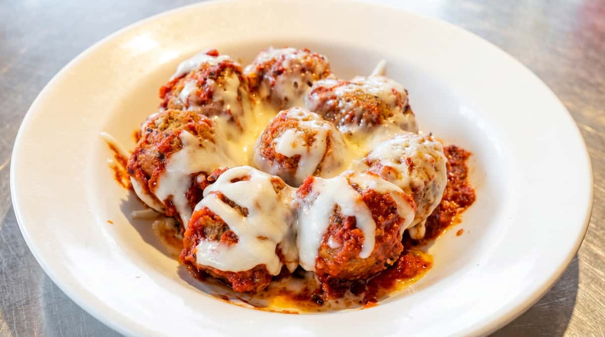 Meatballs covered in red sauce and melted white cheese on a white plate.