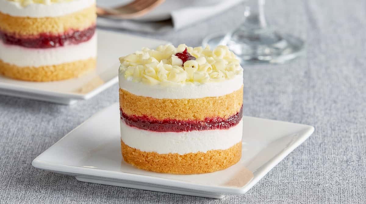 Miniature layered cake with vanilla sponge, white cream, and red fruit filling, topped with white chocolate curls.