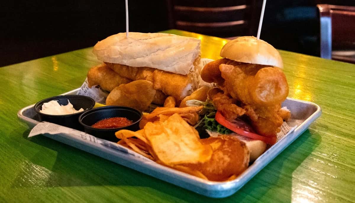 Two crispy fish sandwiches served with house-made chips, tartar sauce, and cocktail sauce on a metal tray.