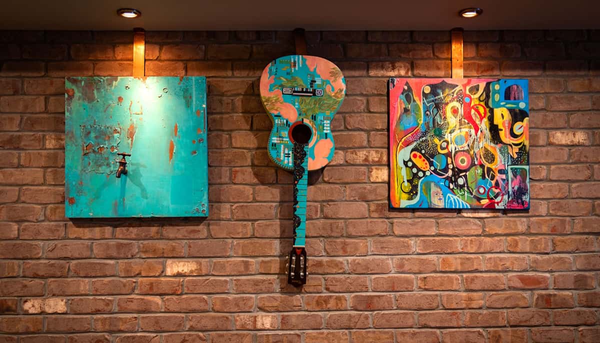 Colorful guitar and abstract paintings displayed on a brick wall under warm lighting.