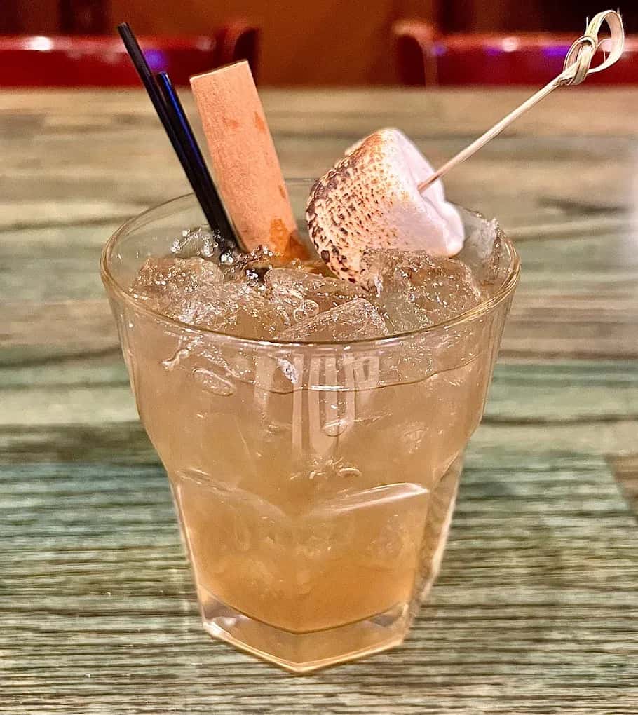 Cocktail with crushed ice, topped with a toasted marshmallow, chocolate stick, and two black straws on a wood-patterned table.