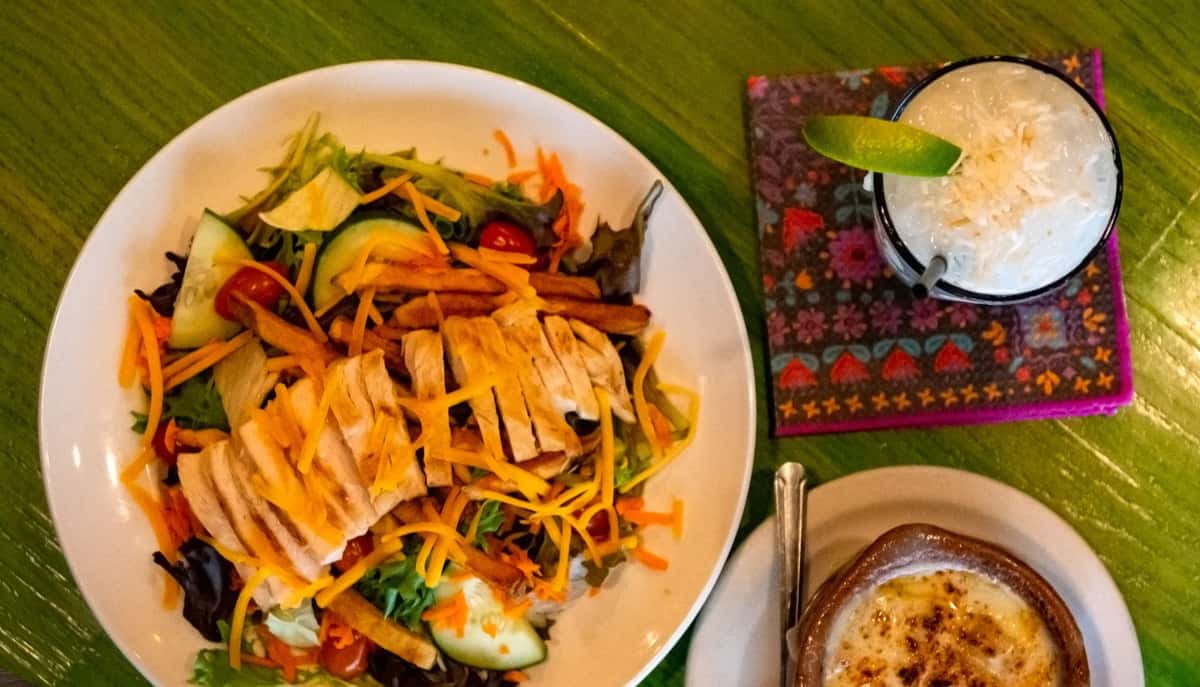 Grilled chicken salad with shredded cheese and veggies, served with French onion soup and a coconut cocktail with a lime wedge.