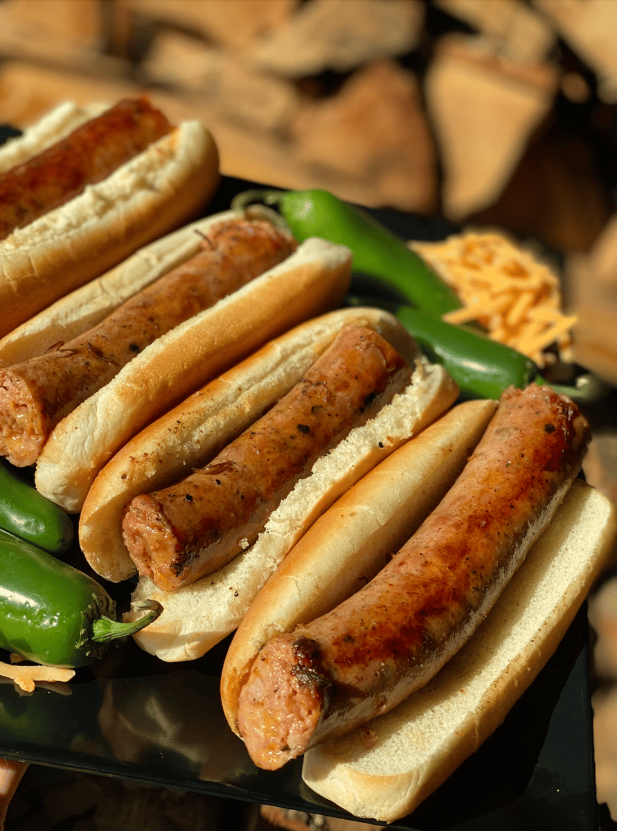 Four J.D.'s Bar-B-Que grilled sausage hot links in buns, plated with bright green jalapeño peppers and shredded cheese, set against a blurred wood background.