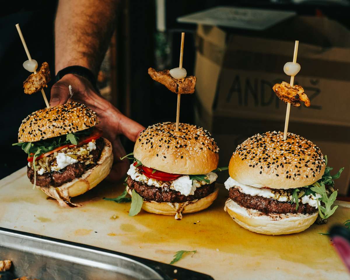 Three gourmet burgers with sesame buns, arugula, tomato, and cheese, topped with skewered grilled meat and onion.