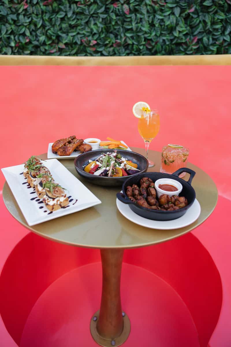 Assorted restaurant appetizers, crispy wings, fresh salad, flatbread bites and handcrafted cocktails served on a vibrant patio table with modern outdoor dining ambiance.
