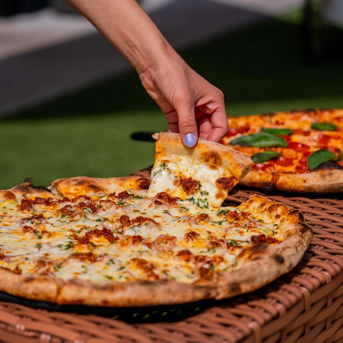 Hand lifting a slice from a freshly baked gourmet cheese pizza with a crispy wood-fired crust, showcasing melted cheese and artisan pizza craftsmanship.