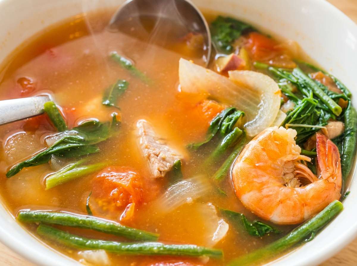 a steaming bowl of shrimp soup with fresh vegetables, including green beans and leafy greens, in a rich, flavorful broth being served with a ladle.