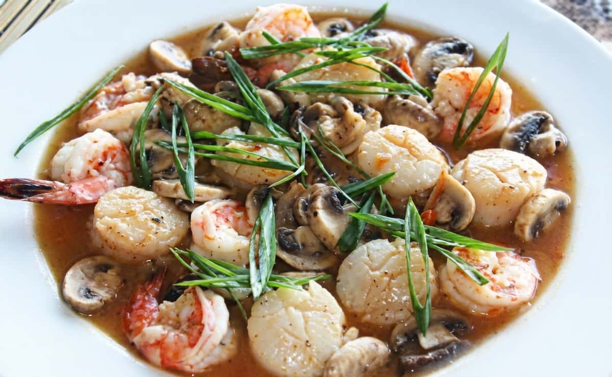 A flavorful seafood dish featuring shrimp, scallops, and mushrooms sautéed in a savory sauce and topped with fresh green onions, served sizzling in a hot plate.