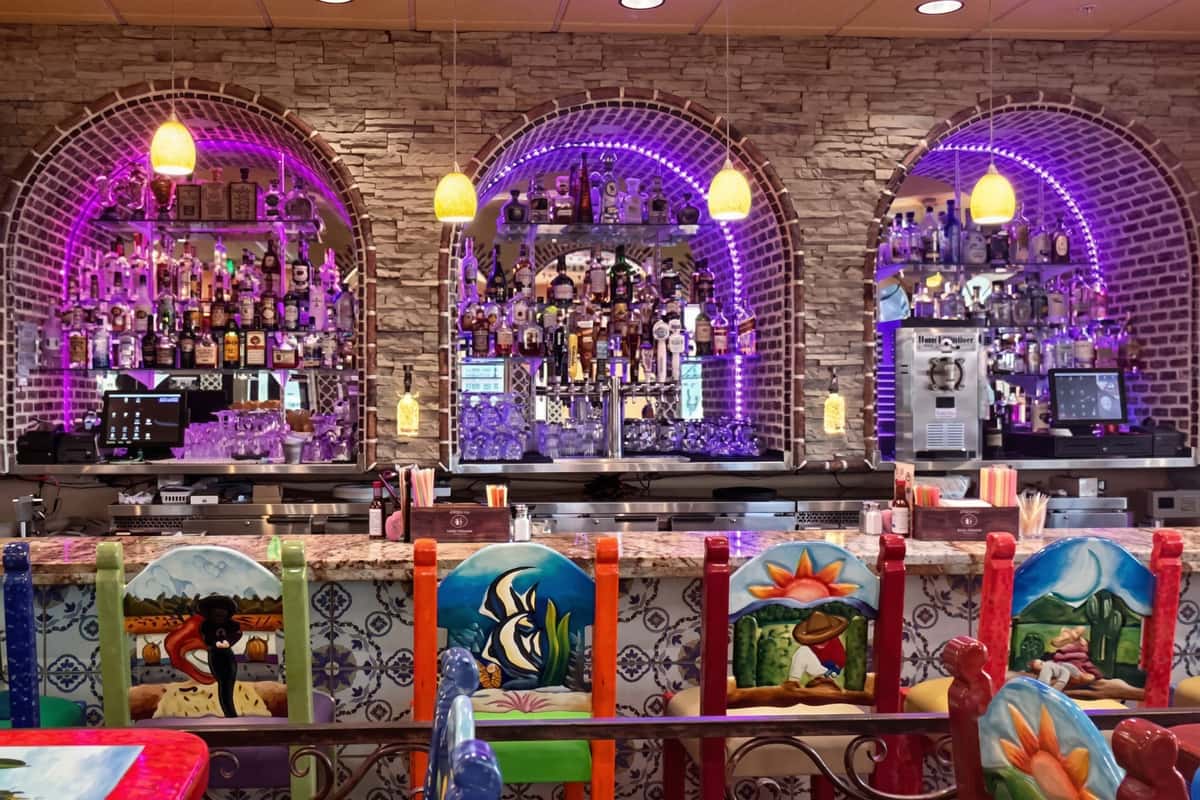 A lively bar area with vibrant purple lighting, showcasing a wide selection of bottles displayed within an arched brick alcove. Colorful decorative panels and traditional Mexican design elements add character, creating a festive and energetic atmosphere.