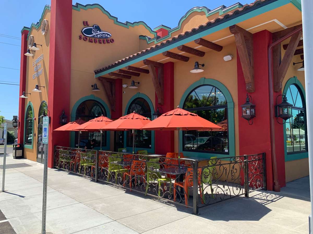 A vibrant Mexican restaurant exterior featuring warm red stucco walls, decorative trim, and a tiled roof. The outdoor patio is lined with colorful railings and shaded by bright umbrellas, creating a lively and inviting space for open-air dining.