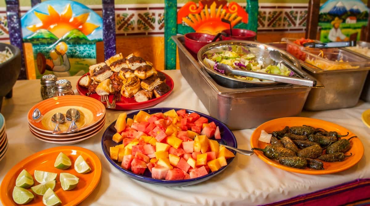 A colorful buffet spread featuring fresh-cut fruit in the foreground, alongside a variety of Mexican dishes including grilled items and a large tray of prepared food. The setup is vibrant and abundant, with warm tones, traditional decor, and a welcoming, festive atmosphere perfect for group dining or catering.