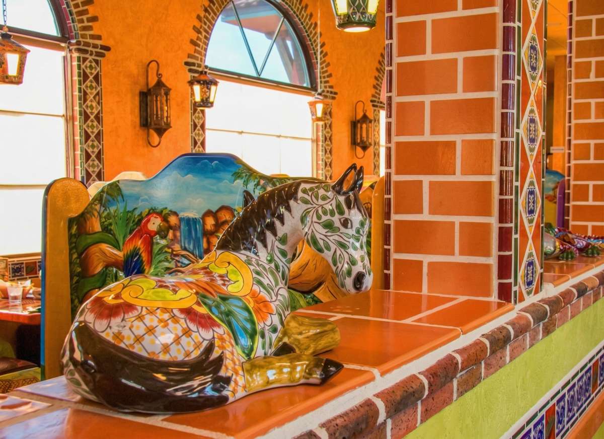 A charming decorative corner featuring a colorful tiled mural of a traditional Mexican village scene. Set against warm terracotta walls and brick details, the space includes handcrafted pottery and vibrant accents, showcasing authentic artistry and cultural character.