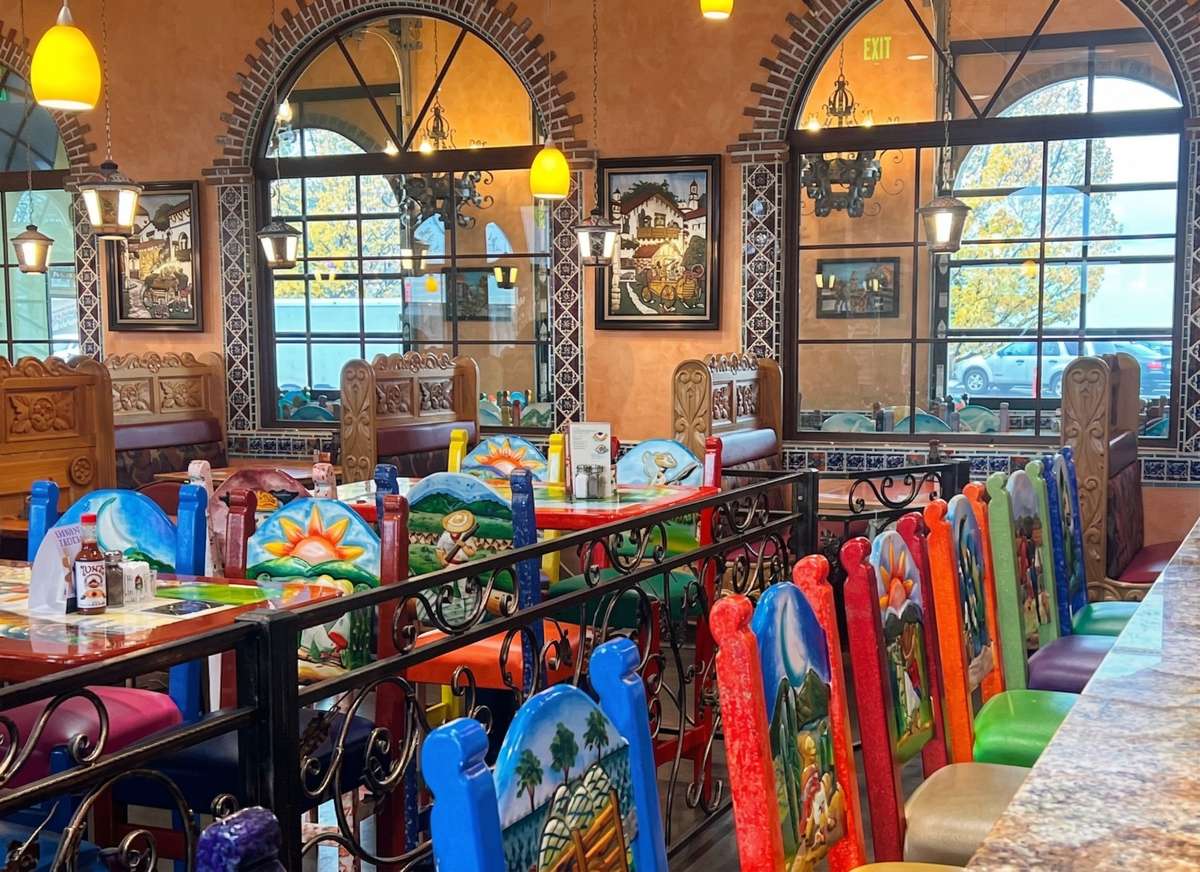 A vibrant and cozy restaurant interior filled with colorful hand-painted chairs and rustic wooden tables. Large arched windows framed with brick and decorative tiles let in natural light, while warm pendant lighting and traditional artwork enhance the inviting, authentic Mexican atmosphere.