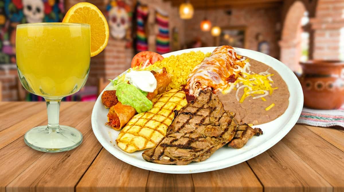 A hearty Mexican-style plate featuring sliced grilled steak, seasoned rice topped with shredded carrots, charred corn on the cob, and a side of creamy guacamole. The dish is vibrant and generous, served on a rustic wooden table with a drink in the background, evoking a warm, casual dining experience.