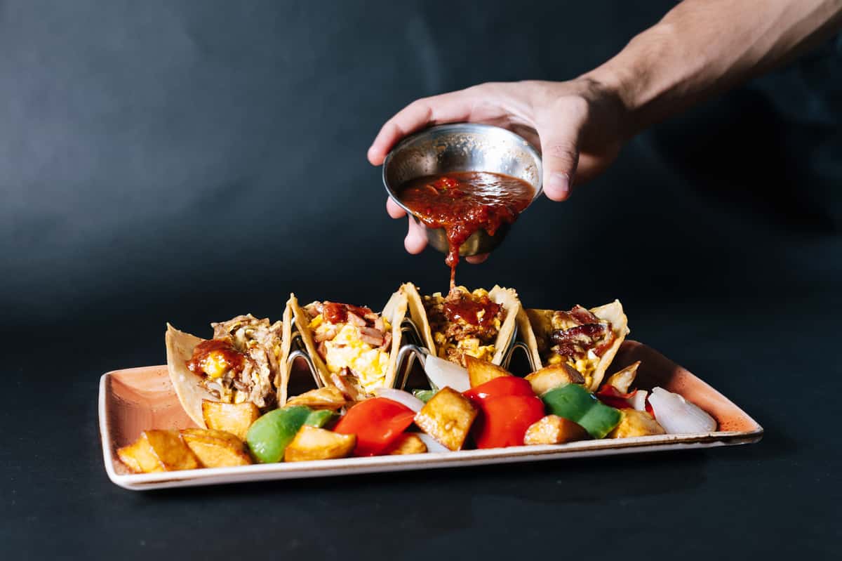 breakfast tacos topped with eggs, cheese, and fresh toppings with salsa being poured over on a platter