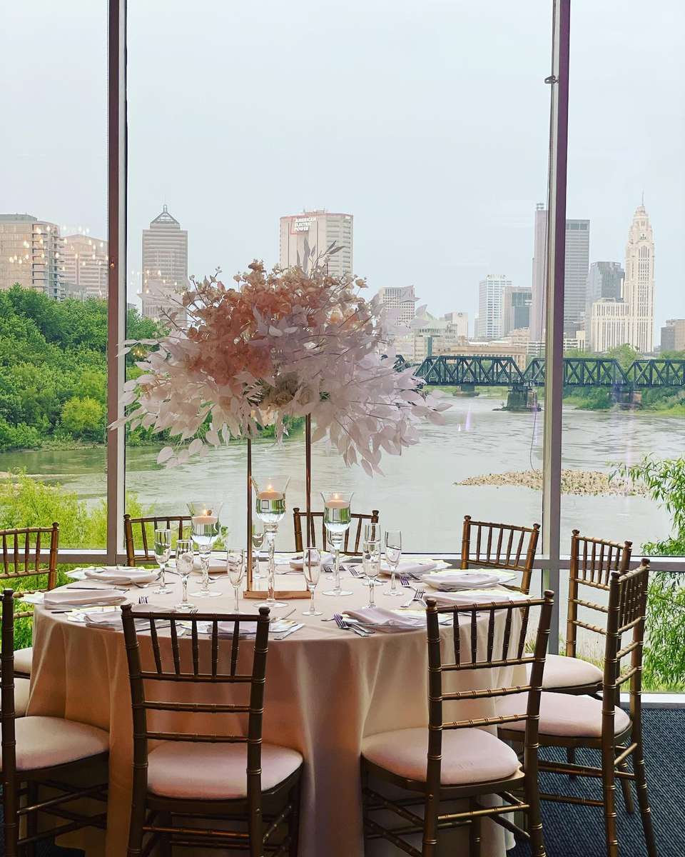 Indoor event space overlooking a river with a skyline view of Columbus. Event tables are customized with cream linens and light pink decor.