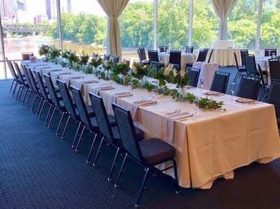 The Buckeye Room in Columbus Ohio, elegantly arranged for a wedding, with a long table set for a private event. The table is adorned with green and white floral centerpieces and flanked by stylish black chairs, all under soft ambient lighting with a view of the waterfront through draped windows