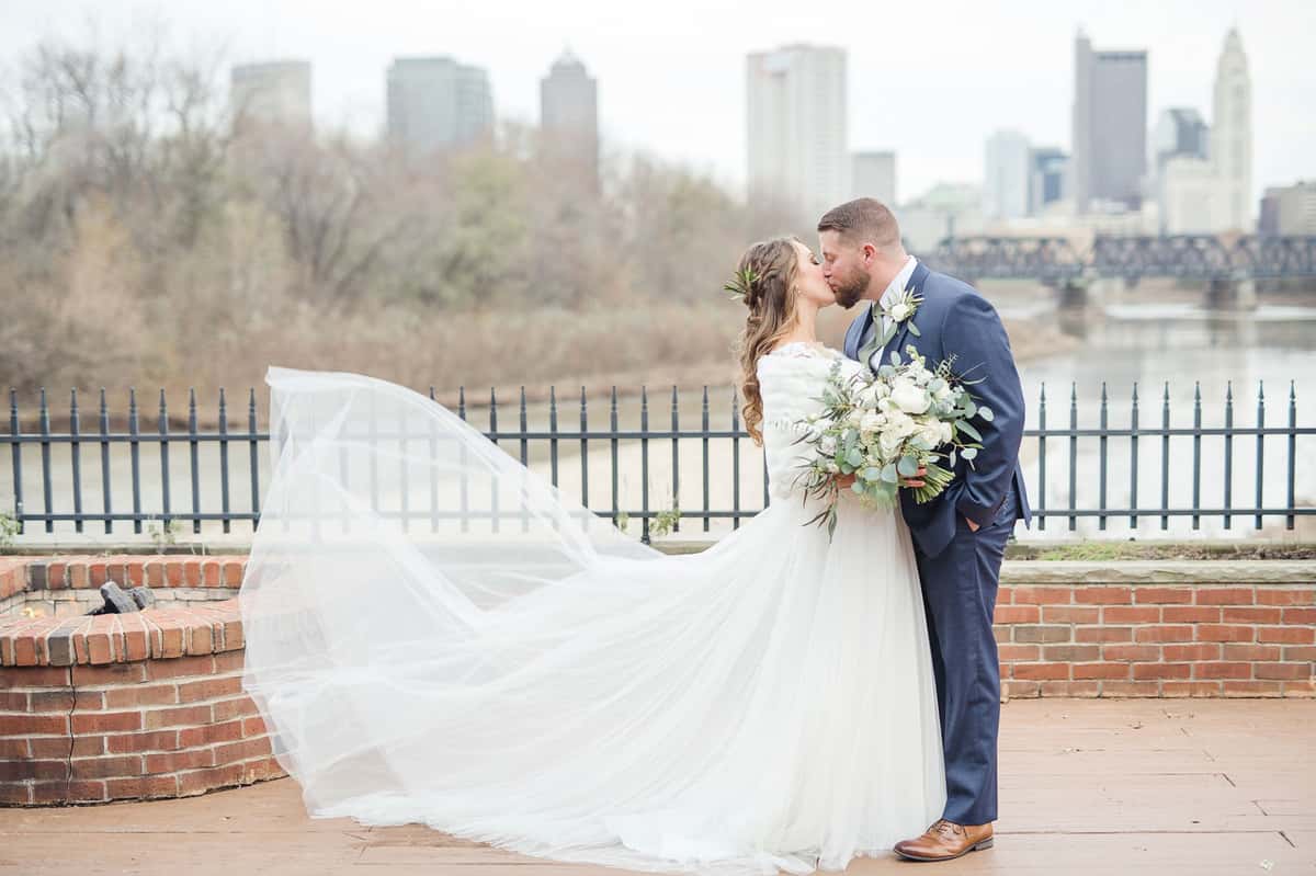 Beautiful bridde kissing her groom on a bridge with a city skyline view. Weddings and wedding receptions in Columbus, OH