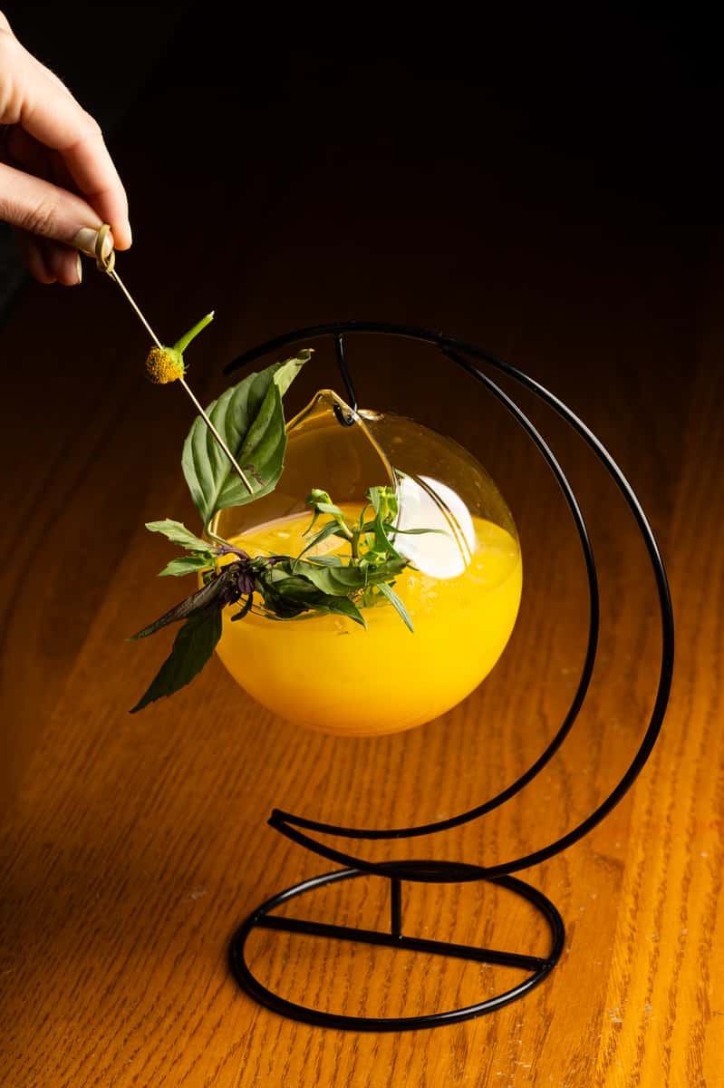 Modern yellow craft cocktail with fresh herb garnish served in a unique suspended glass presentation at an upscale cocktail lounge