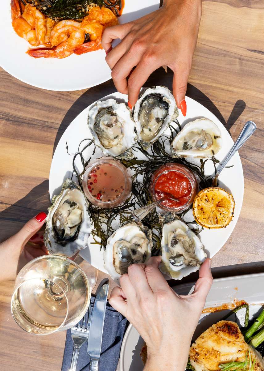 Fresh oysters on the half shell served with cocktail sauce, mignonette, grilled lemon, and seaweed garnish on a seafood restaurant table with white wine and shared plates