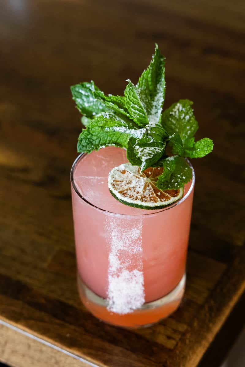 Refreshing pink craft cocktail with fresh mint garnish and dried lime served on a rustic wooden bar table in an upscale restaurant lounge