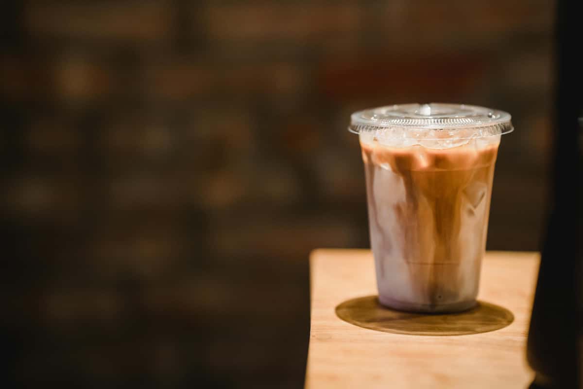 Iced coffee with milk swirls in a clear plastic cup and lid, placed on a wooden surface against a blurred brick background.