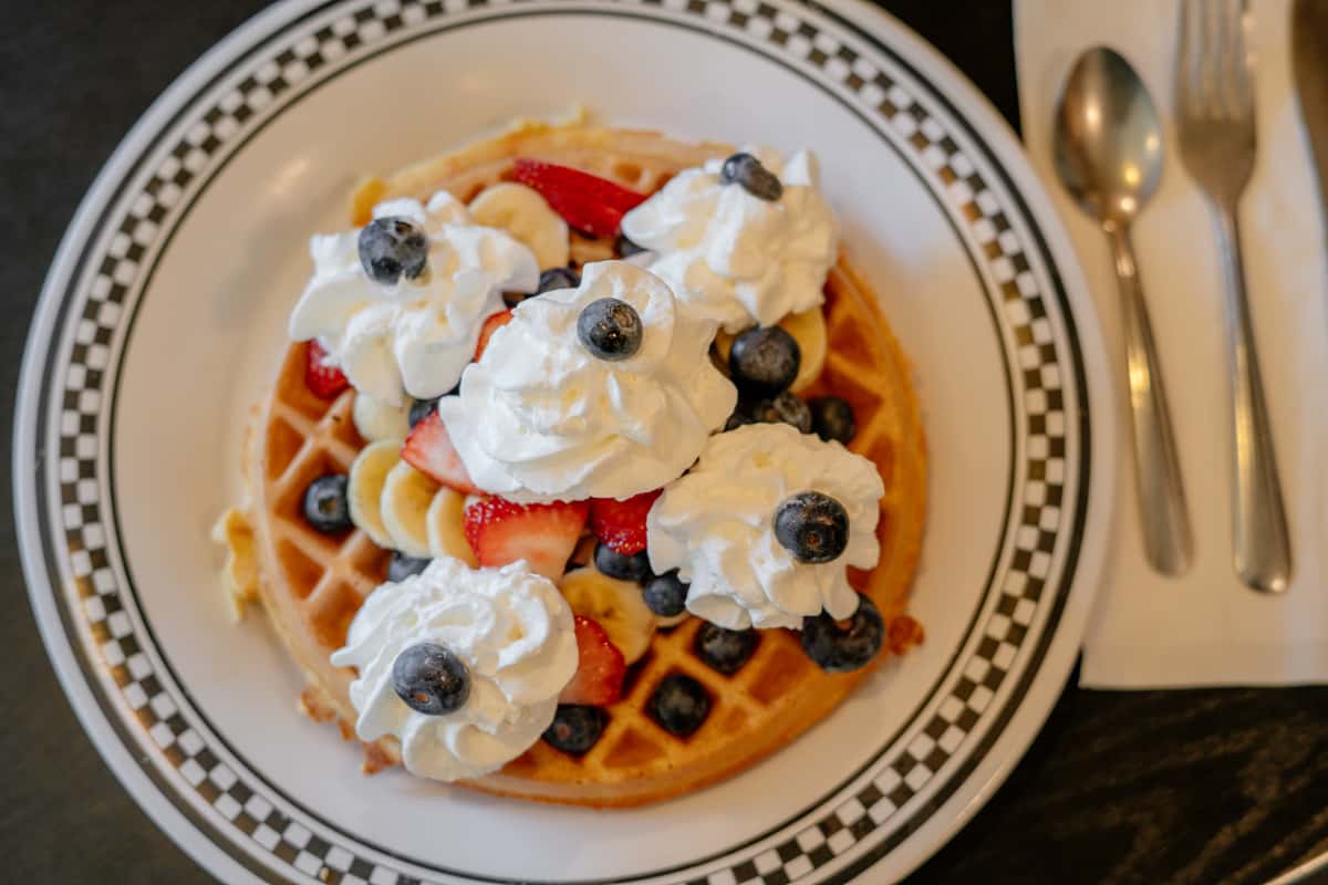 A golden Belgian waffle topped with fresh strawberries, blueberries, and banana slices, finished with generous whipped cream and a light drizzle of syrup. Served on a classic diner-style plate, this colorful and indulgent dish highlights a sweet, fresh breakfast favorite, perfect for a comforting morning treat at a cozy café.