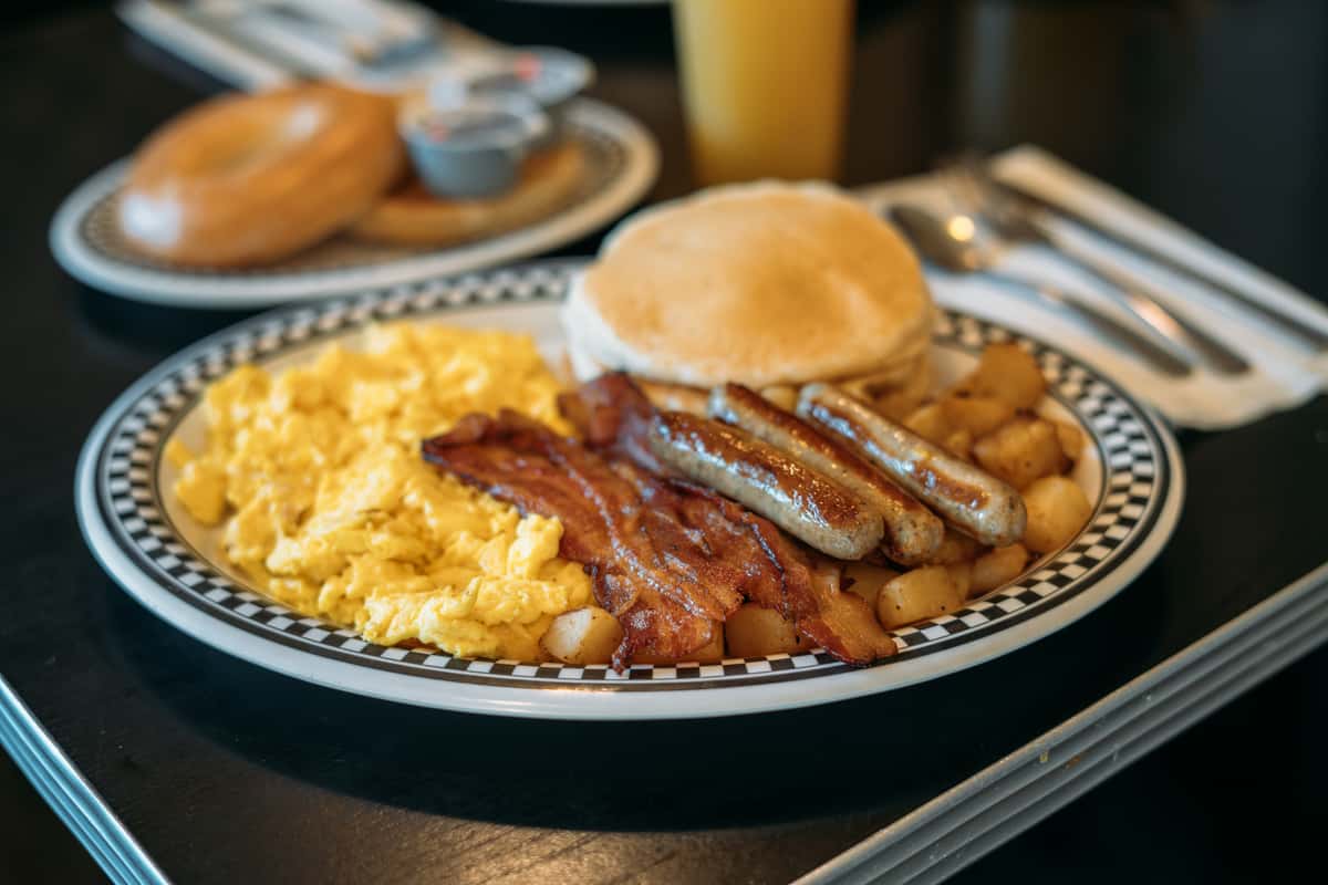 A hearty breakfast plate featuring scrambled eggs, crispy bacon, breakfast sausage, baked beans, and a toasted English muffin, served on a classic diner-style plate. In the background, a glass of orange juice and breakfast sides add to the warm, comforting café atmosphere. The dish highlights a traditional homestyle breakfast, perfect for starting the day with satisfying flavors at a cozy neighborhood café like Mel's Commonwealth Cafe.