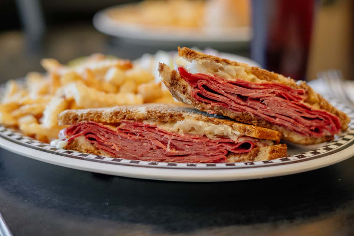 A hearty stacked deli sandwich layered with thinly sliced corned beef, melted cheese, and toasted rye bread, cut to reveal the rich, savory filling. Served alongside a portion of crispy golden French fries, this classic diner-style meal delivers bold flavor and satisfying comfort—perfect for a filling lunch at a neighborhood café.
