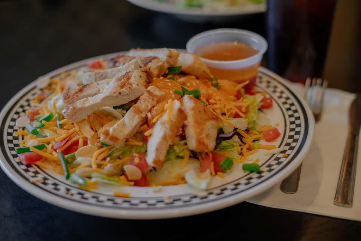 A colorful Southwest-style breakfast plate topped with crispy tortilla strips and sliced grilled chicken, served over a hearty mix of eggs, fresh vegetables, and melted cheese. Finished with chopped herbs and accompanied by a side of salsa, this dish offers a bold, savory blend of flavors, perfect for a satisfying and flavorful café breakfast or brunch.