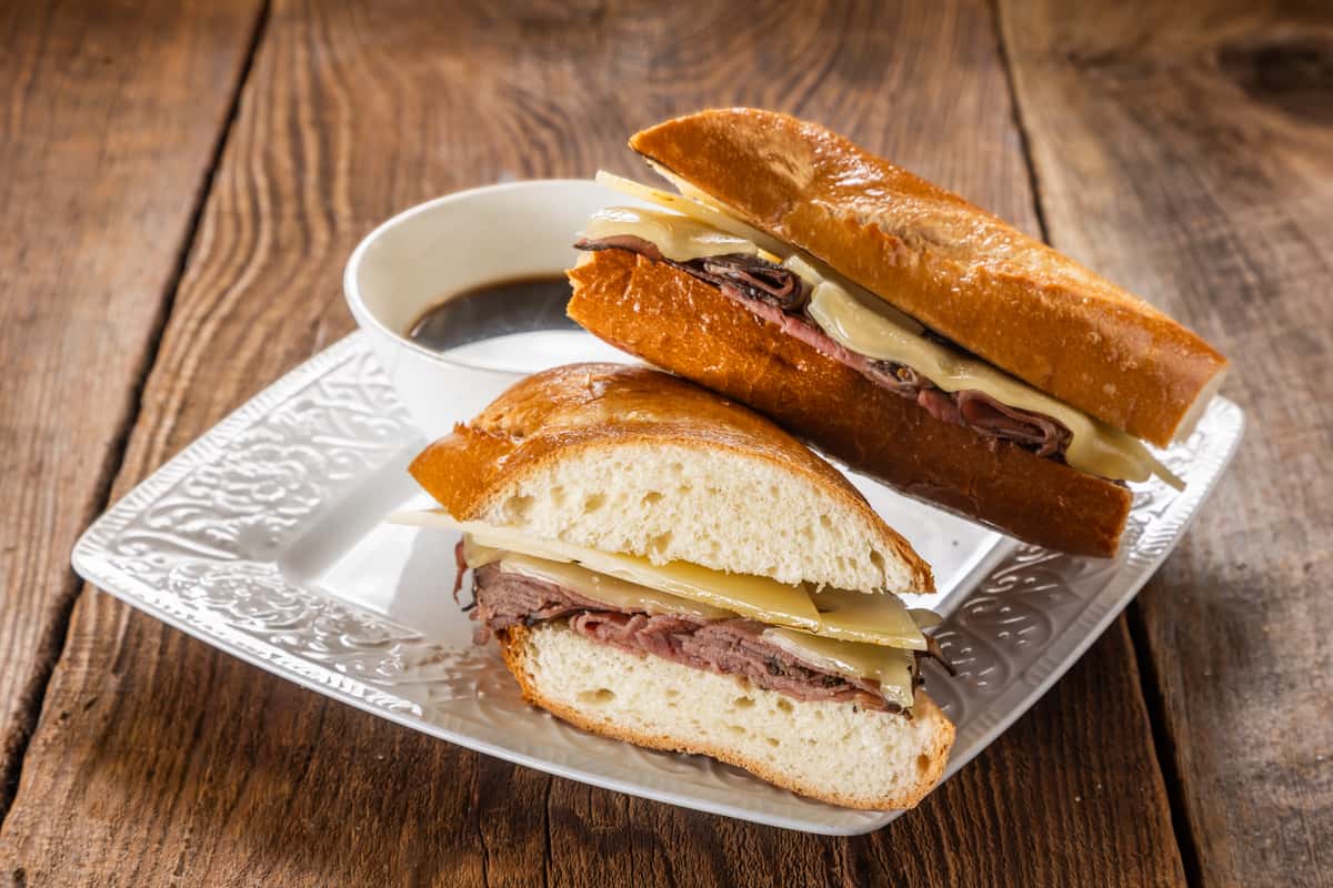 Roast beef and Swiss cheese sandwich on toasted French bread served with a side of savory au jus dipping sauce on a white plate over a rustic wooden table.