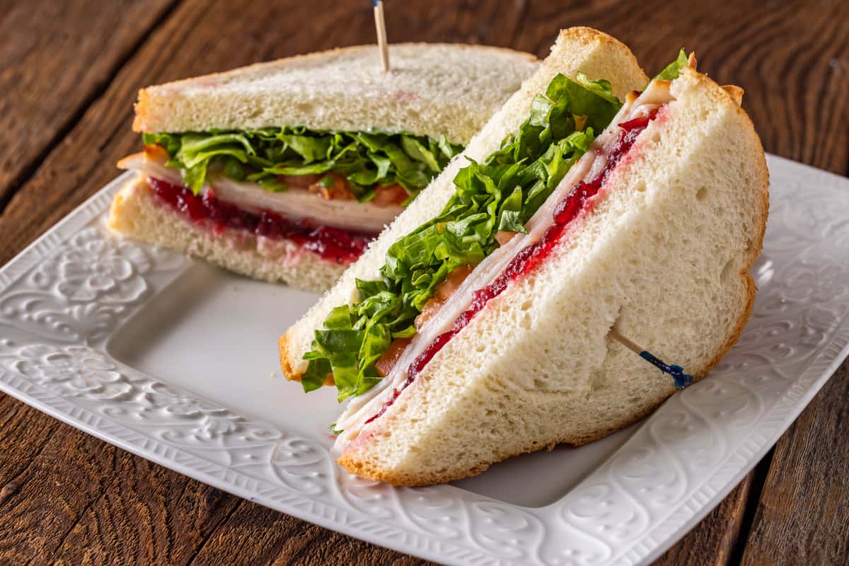 Turkey sandwich on soft white bread with cranberry spread, fresh leafy greens, and sliced turkey breast served on a white plate over a rustic wooden table.
