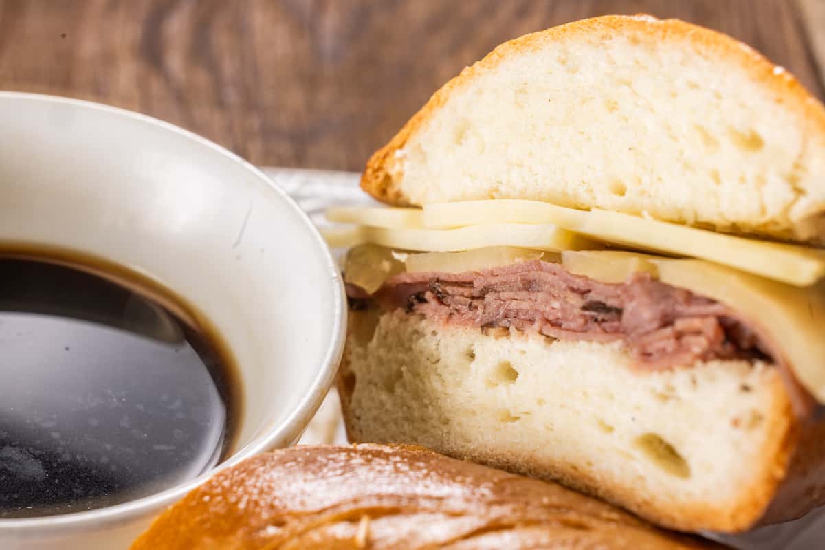 Roast beef and Swiss cheese sandwich on rustic artisan bread served with a side of savory au jus dipping sauce, photographed on a wooden table for a classic deli-style presentation.