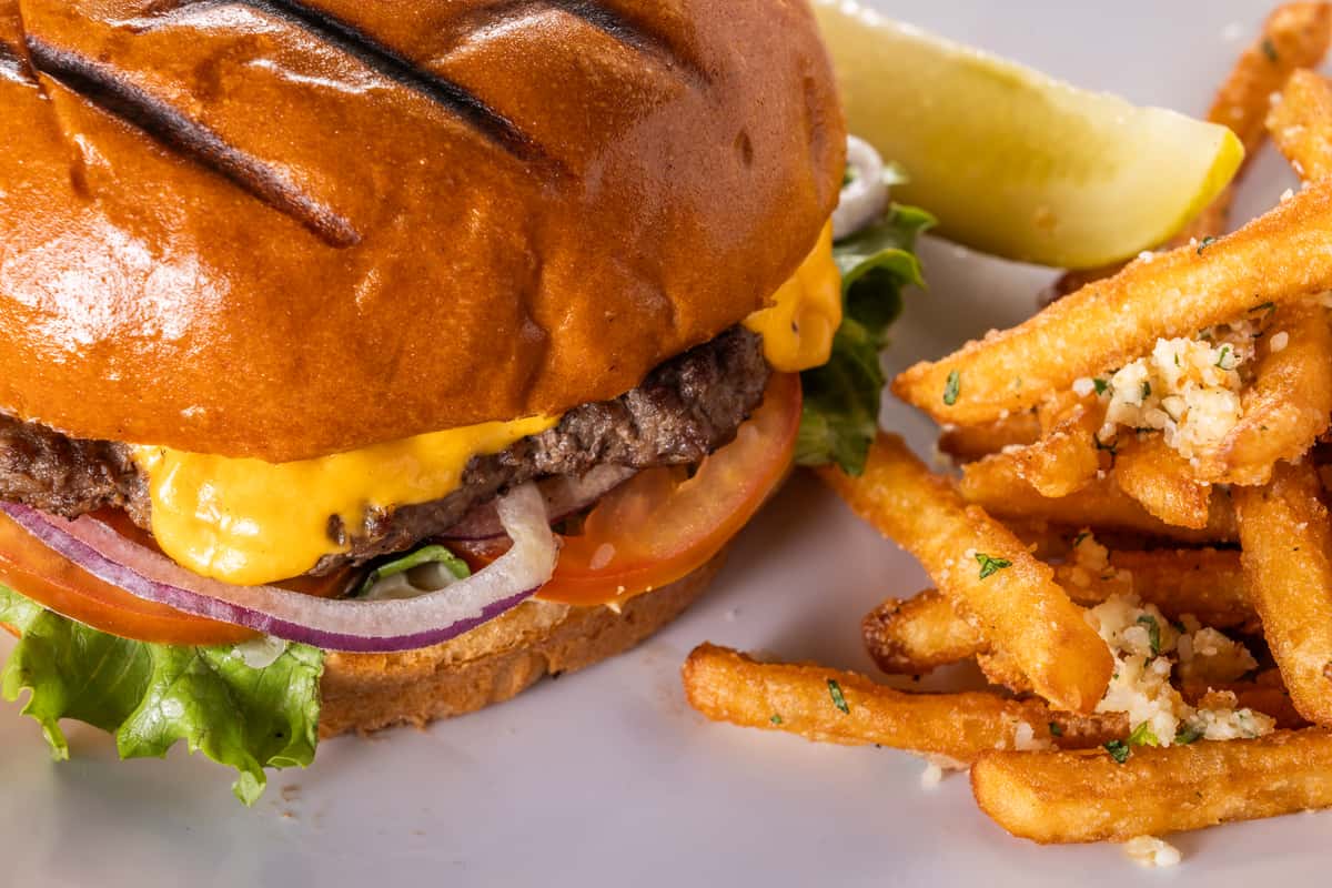 Grilled cheeseburger with melted cheddar, lettuce, tomato, and red onion on a toasted brioche bun served with crispy garlic-seasoned French fries and a pickle wedge on a white plate.