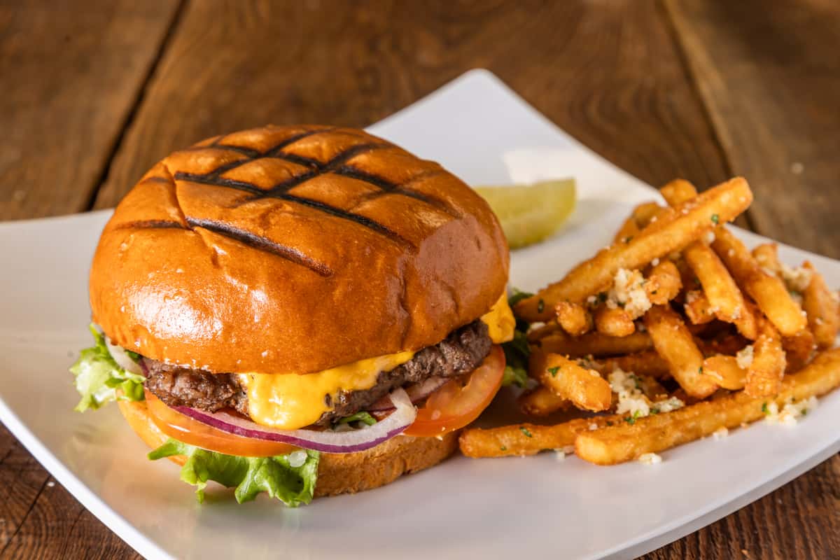 Grilled cheeseburger with melted cheddar, lettuce, tomato, and red onion on a toasted brioche bun, served with crispy seasoned French fries on a white plate over a rustic wooden table.