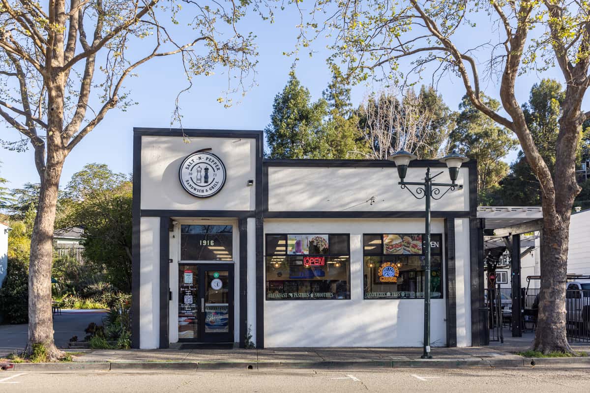 Restaurant storefront with modern white exterior and black trim, featuring large windows, an illuminated open sign, and trees lining the street in a neighborhood dining district.
