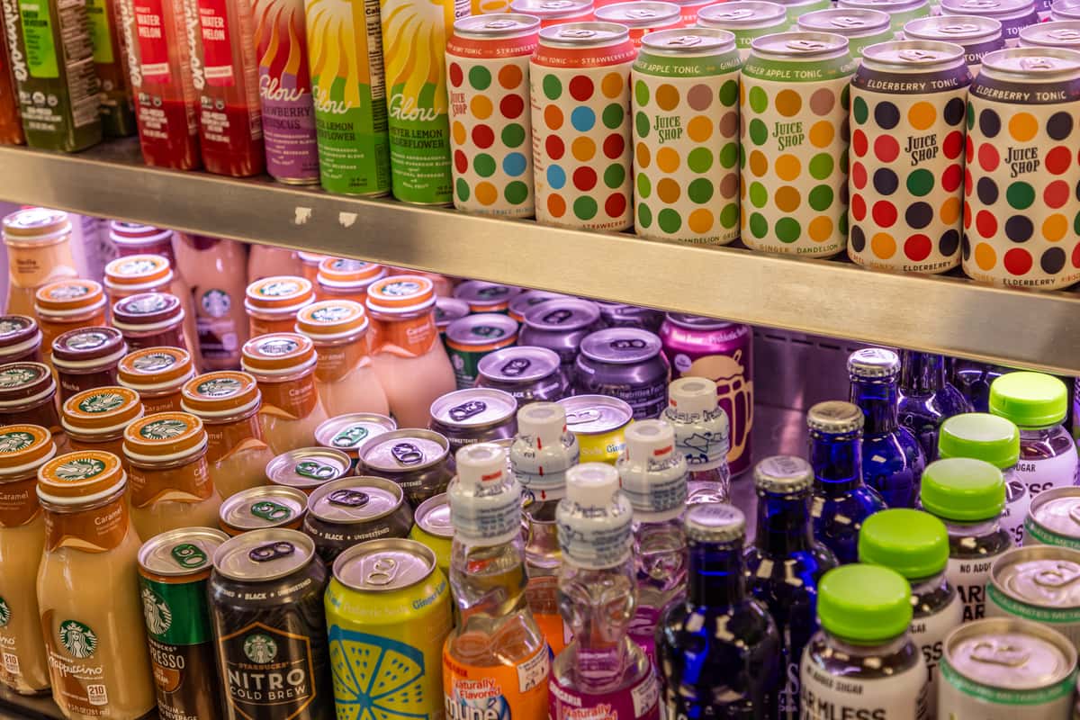 Refrigerated beverage display stocked with colorful canned drinks, bottled juices, kombucha, sparkling tonics, and Starbucks cold brew in a modern restaurant grab-and-go cooler.