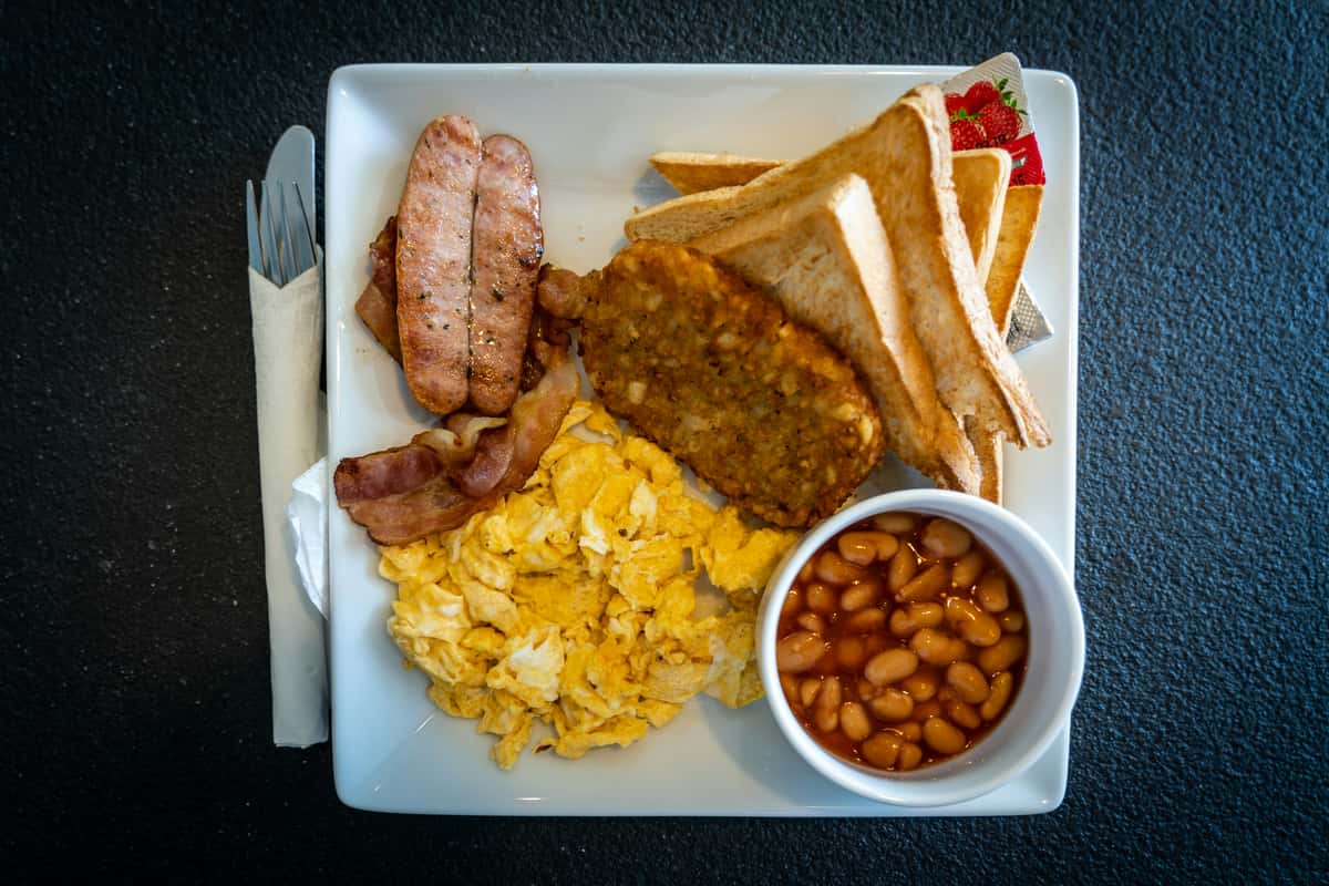 Scrambled eggs, bacon, sausages, hash browns, toast, and baked beans served on a square white plate.