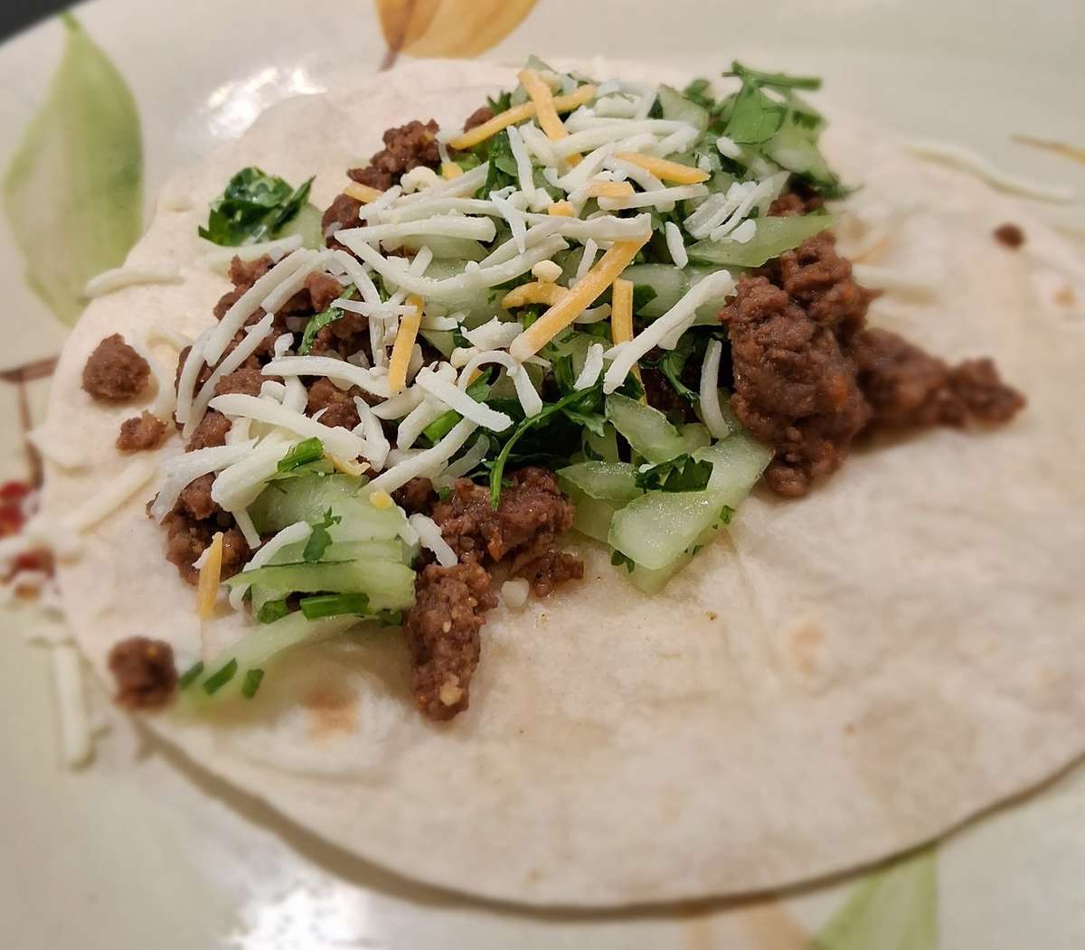 Soft taco with seasoned ground beef, shredded cheese, cucumber slices, and fresh cilantro on a flour tortilla.