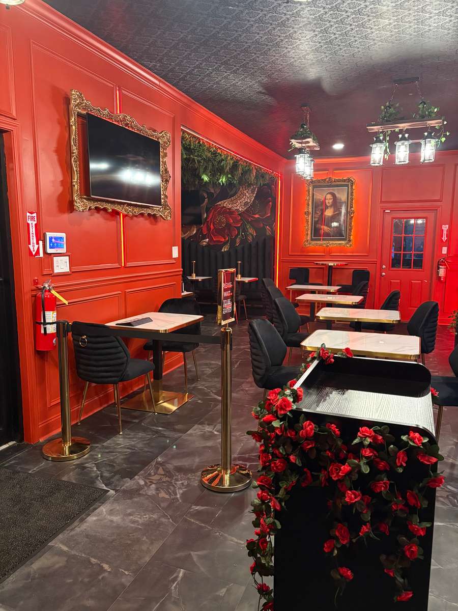 Interior of a restaurant with red walls, featuring a TV, vintage-style portraits, a flower-decorated counter, and elegant black chairs with gold accents.