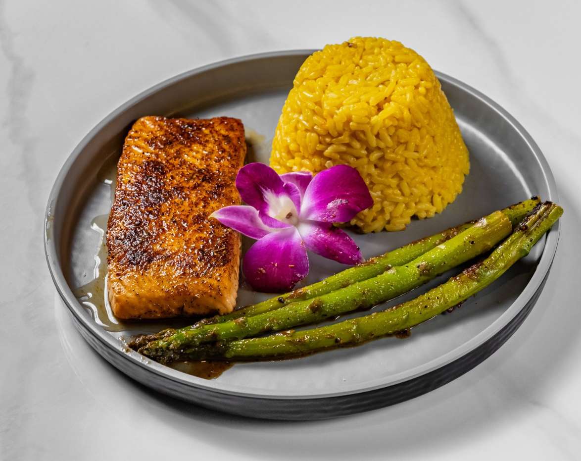 "Grilled salmon fillet served with a mound of yellow rice, asparagus, and a purple orchid garnish on a gray plate."