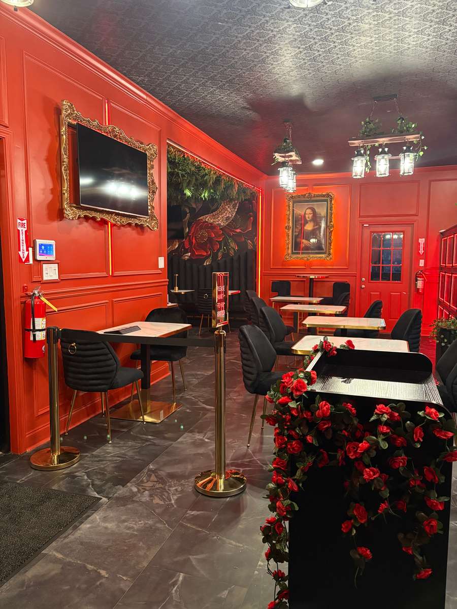 Interior of a restaurant with red walls, featuring a TV, vintage-style portraits, a flower-decorated counter, and elegant black chairs with gold accents.