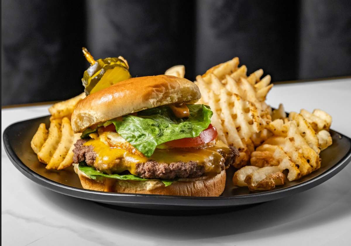 Cheeseburger with lettuce, tomato, and pickles on a toasted bun, served with crispy waffle fries on a black plate.