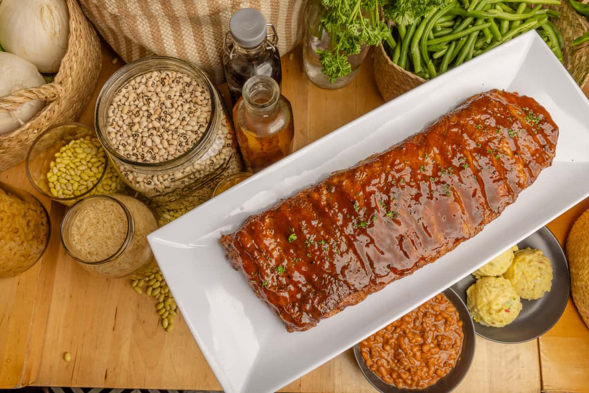 Barbecue glazed rack of ribs on white platter with cornbread, baked beans, rice, and fresh vegetables