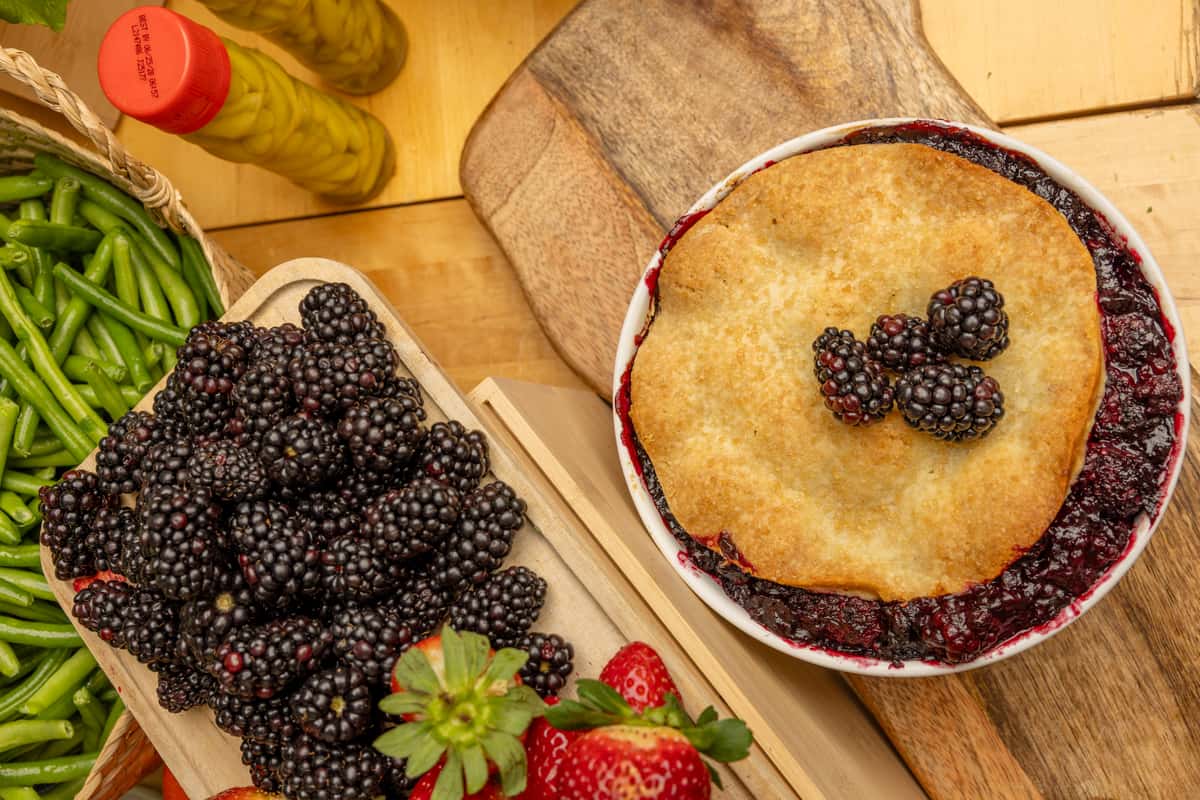 Overhead view of blackberry cobbler with golden crust, fresh blackberries, green beans, and strawberries on wooden boards.