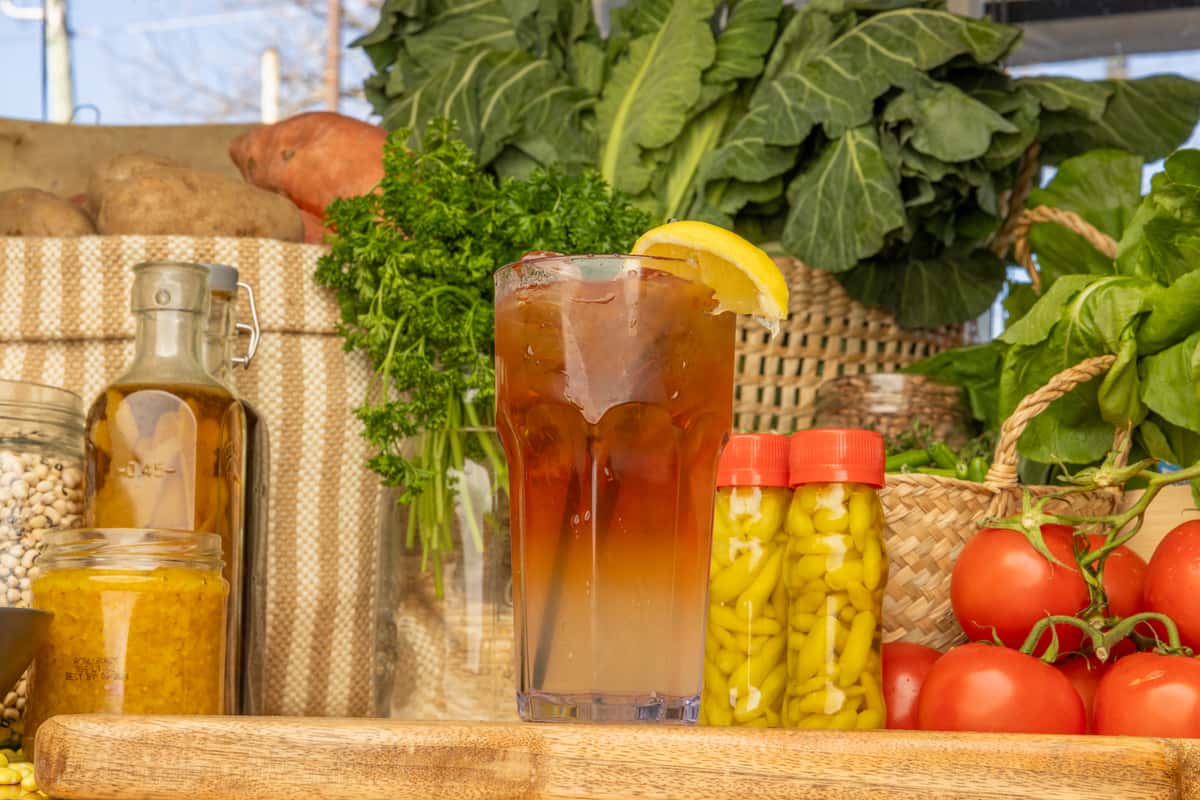 Tall glass of iced tea with lemon slice, surrounded by collard greens, tomatoes, pickled peppers, and pantry items.