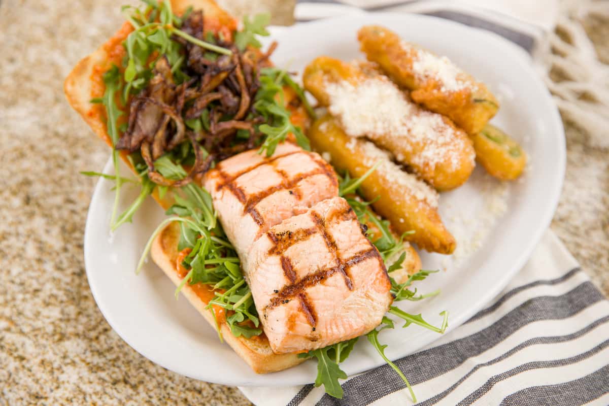 A grilled fish fillet with distinct char marks is served alongside a fresh green salad and a side of golden, crispy breaded shrimp. The dish is plated on a white dish with a light drizzle of sauce, combining a healthy seafood option with a satisfying, crunchy element.