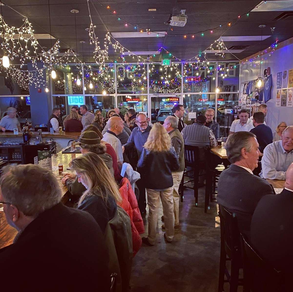 Lively bar filled with patrons enjoying drinks and conversation under festive string lights and colorful decor.