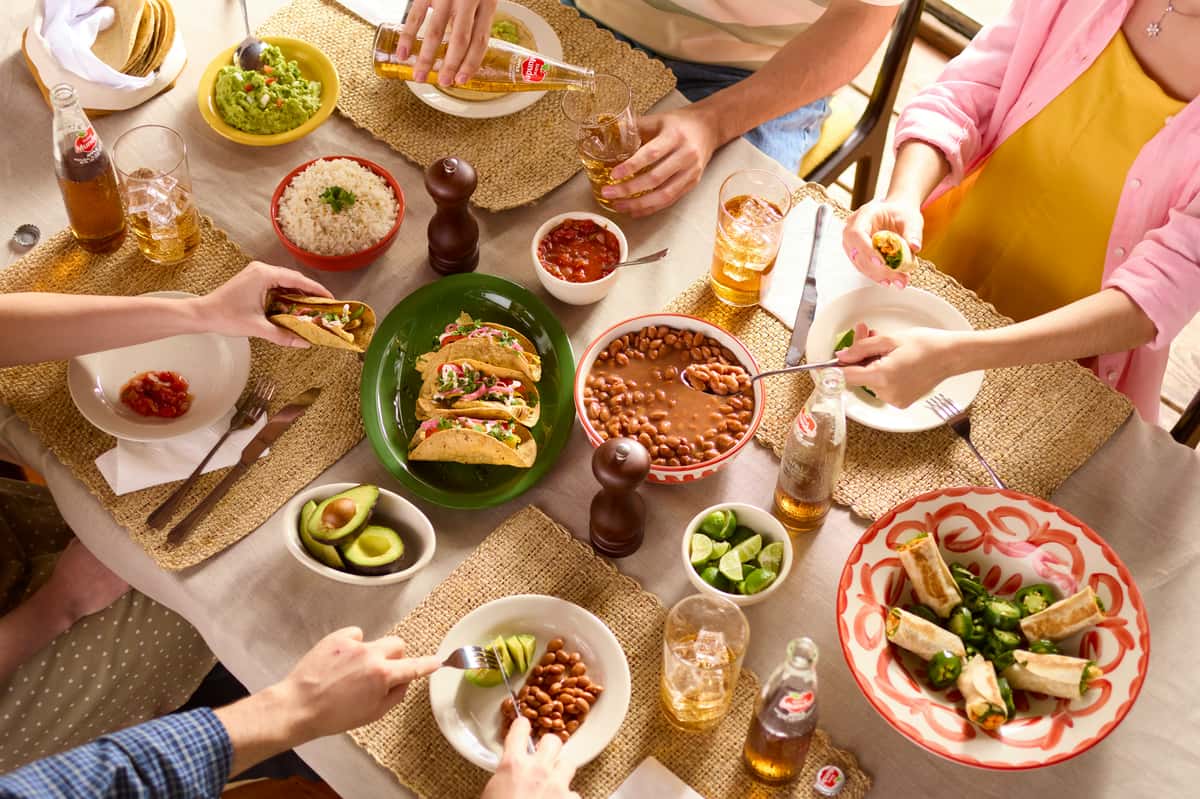 Overhead view of a shared Mexican meal with tacos, beans, rice, guacamole, and drinks—family-style dining with fresh ingredients.